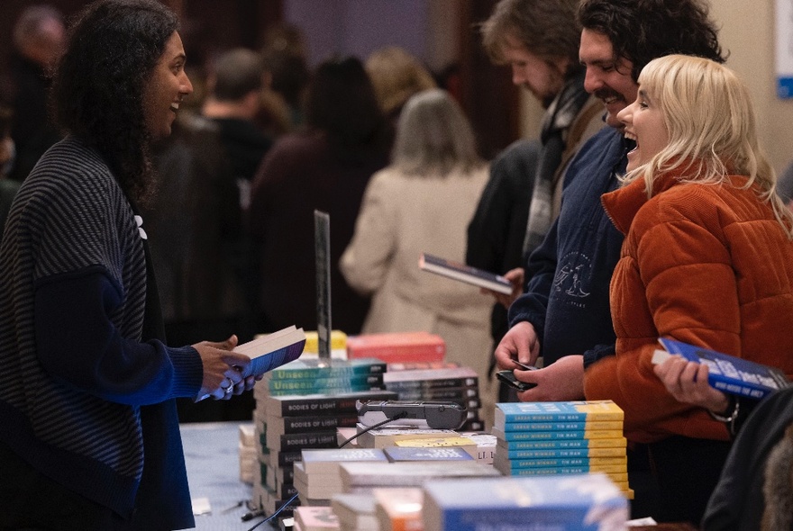 People conversing at a book signing with piles of books on a long table.