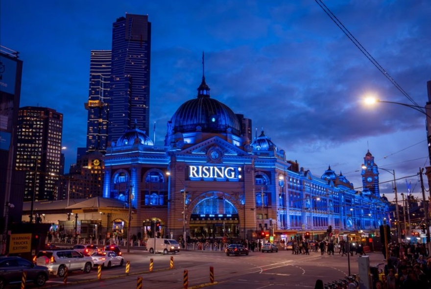 Melbourne cit intersection showing the exterior of Flinders Street Station building illuminated in blue lights and with the words RISING displayed under the dome at the main entrance; dusk, skyscrapers in the background, pedestrians and some cars in the foreground.