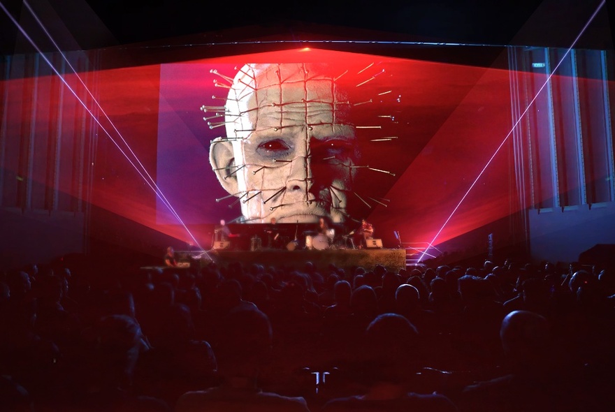 Audience seated in a dark theatre, facing a band playing live on red-lit stage, while above the stage a screen projects an image of a head with many long nails protruding out of it, laser light beams also projected from the base of the stage to the ceiling.