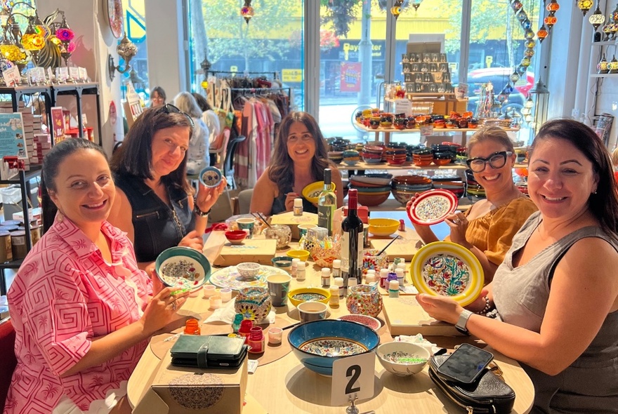 A group of people seated at a crafts table covered in pots of paint and crafts accessories, each holding their painted bowl, with crowded shop displays in the background.