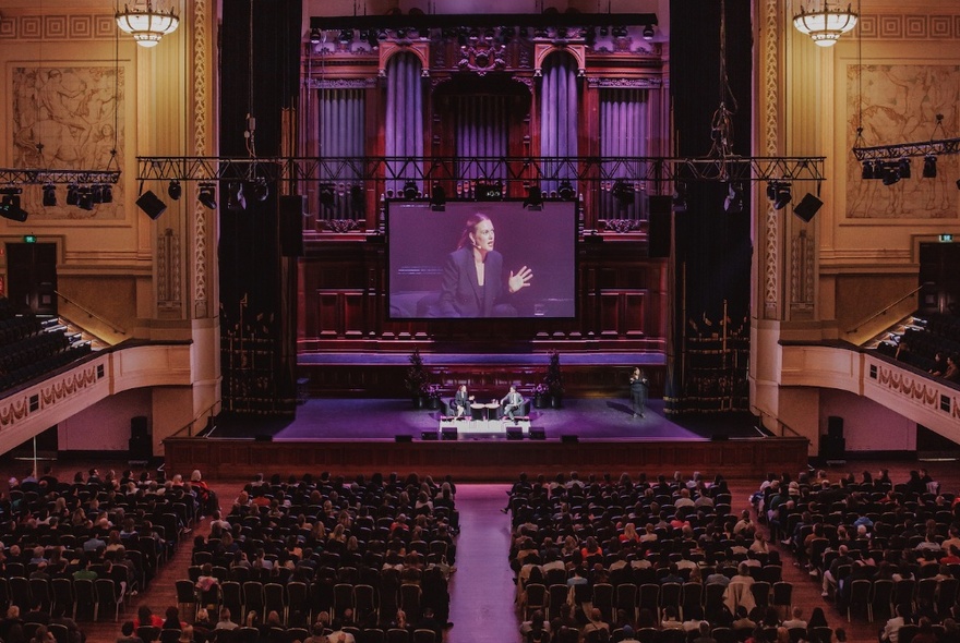 The interior of a large hall with a seated audience and an interview being conducted on stage and also projected on a large screen. 