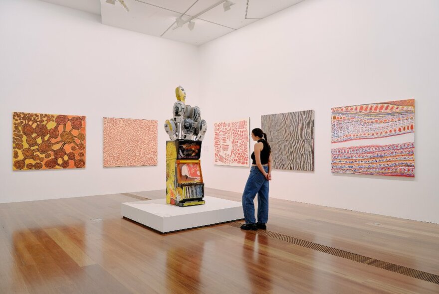 A person looking at Indigenous artwork hanging on the walls of a gallery and a platform displaying a sculpture.
