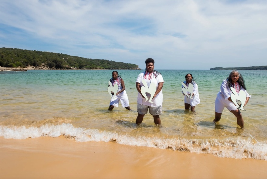 Four dancers performing knee-deep in clear water on a beach.