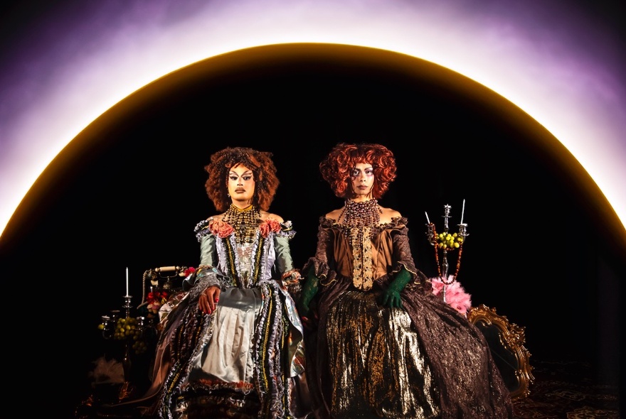 Two drag queens in renaissance inspired dresses in front of a background that resembles a lunar eclipse.