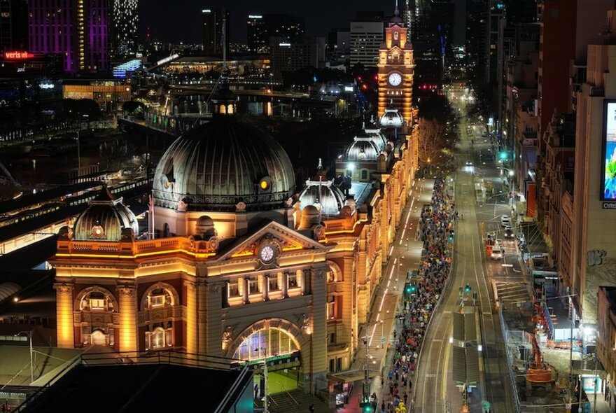 An aerial view looking up Flinders St at night with Flinders Street Station illuminated towards the left of the image. 