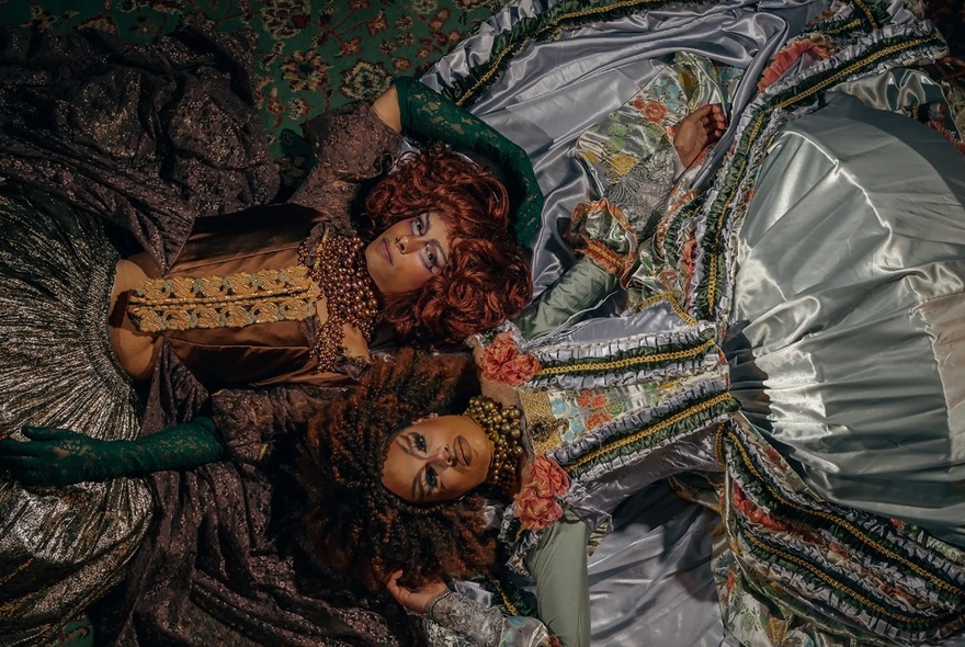 Looking down on two drag queens in renaissance inspired dresses, heads together by in different directions. 