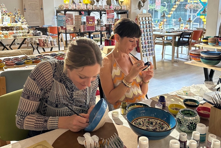 Two people seated at a crafts table, painting bowls with brushes.