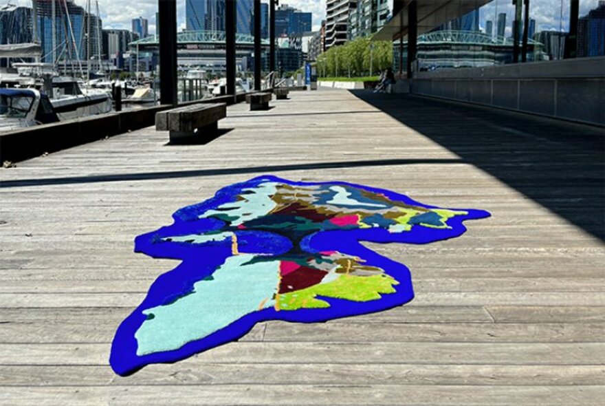 A very brightly coloured rug, in the shape of an island, displayed on a wooden pier.