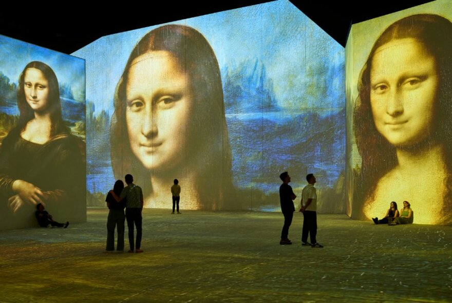 People in a very large space with the Mona Lisa artwork projected on all the wall spaces. 