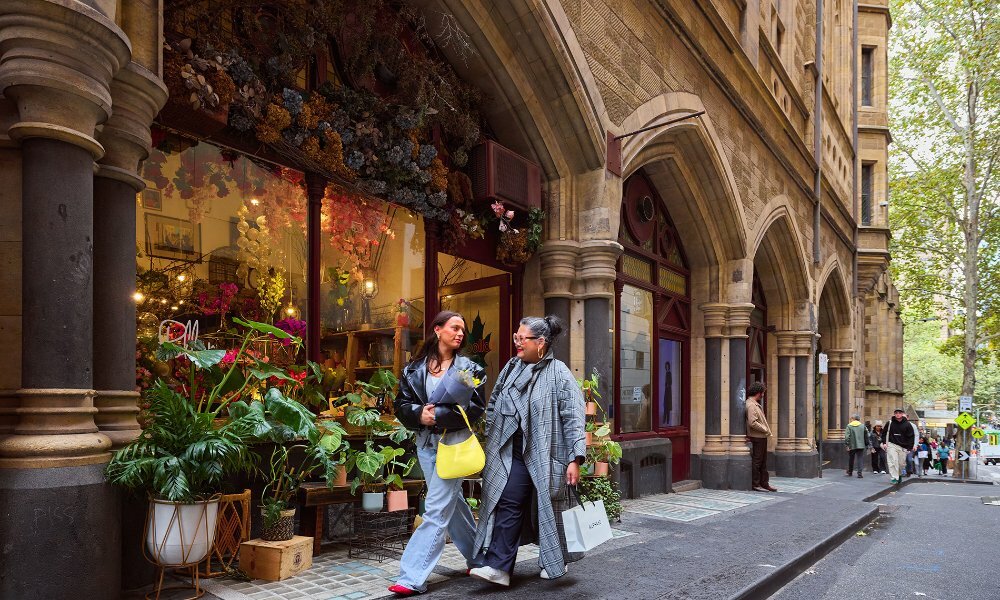 Two women walking past a florist with shopping bags and a bunch of flowers in their arms.