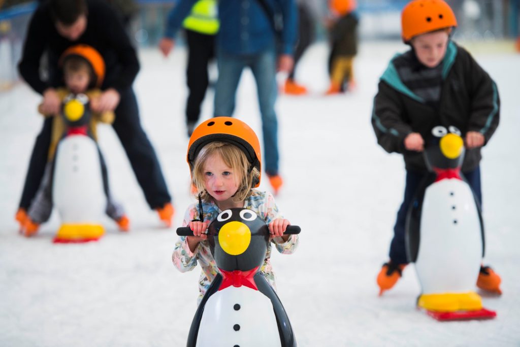 A child with a helmet on pushing a penguin toy and skating on an ice rink